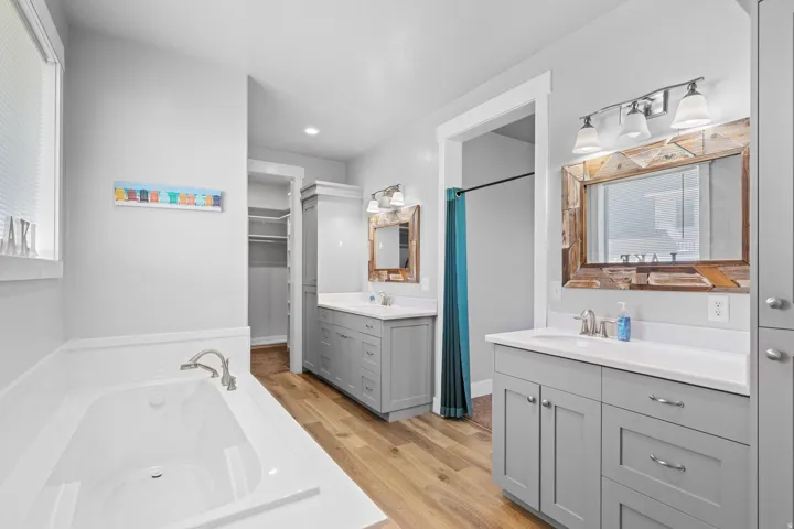 Soaking tub with deck-mounted faucet, dual vanity with shaker-style cabinetry and undermount sinks, wood-finish flooring, and a walk-in closet