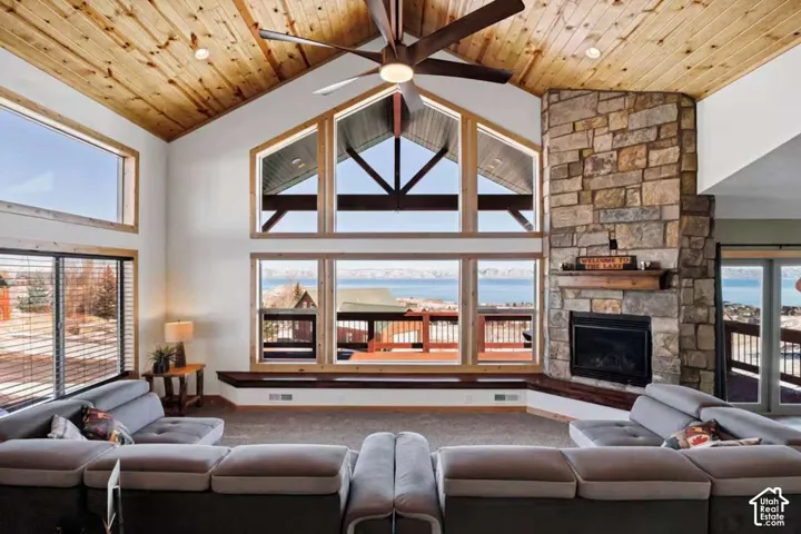 Vaulted ceiling with wood-finish paneling, stone-clad fireplace, large picture windows, sliding glass doors, and a ceiling fan with integrated lighting