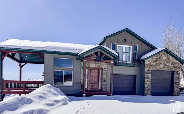 Siding exterior with stone accents, a covered deck, multiple attached garages, a second-story balcony, and a prominent wood-finish front door