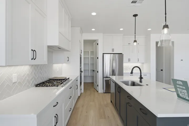 White cabinetry with dark hardware, sleek built-in gas range, herringbone tile backsplash, stainless steel appliances, and a large center island with a built-in sink and pendant lighting