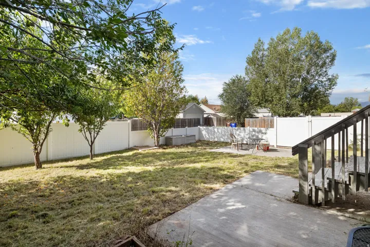 Expansive backyard with a concrete patio, mature trees, and a white privacy fence