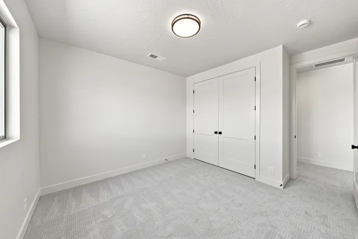 Neutral wall paint, ceiling-mounted light fixture, dual panel closet doors with dark hardware, window with white frame, and textured carpet flooring