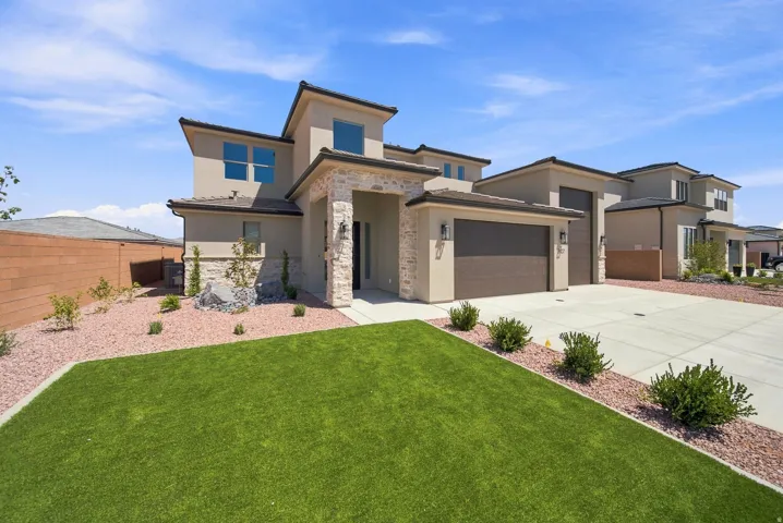Contemporary stucco exterior with stone accents, integrated two-car garage, concrete driveway, manicured lawn, and xeriscaped landscape beds
