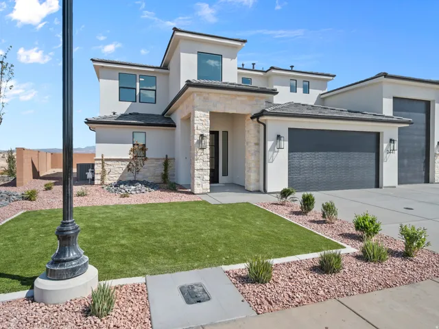 Modern stucco exterior with stone accents, dark garage doors, front entry with transom window, and a low-maintenance landscape featuring artificial turf and decorative rock