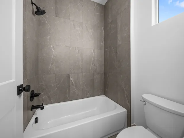 Bathtub with dark gray stone-finish wall tile, matte black shower fixtures, and a white toilet