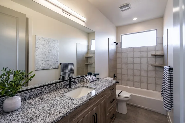 Spa-inspired primary bathroom featuring a dual-sink vanity with speckled stone countertop, abundant natural light from a transom window, and a tile-surround soaking tub with glass enclosure