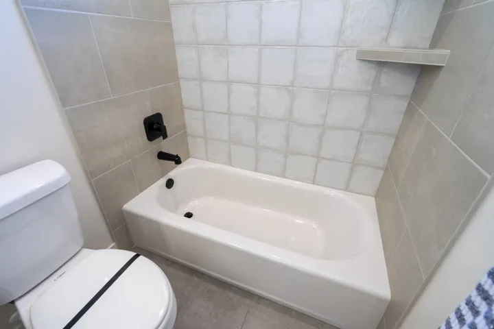 Integrated bathtub with matte black fixtures, light-toned wall tiling, and a tiled shower shelf