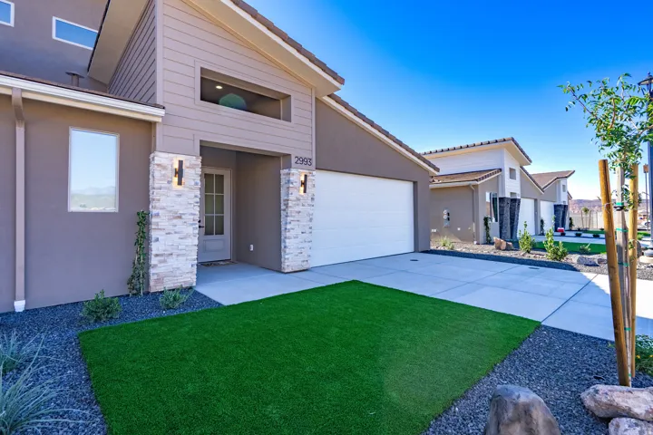 Modern home featuring a white two-car garage with paneled door, contemporary stucco and brick exterior with sconce lighting, manicured front lawn with synthetic turf, decorative gravel landscaping, and clean concrete driveway with integrated hardscaping