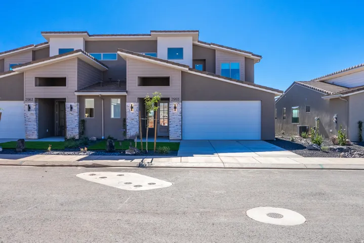 Modern residential home featuring a clean contemporary facade with mixed stone and stucco exterior, dual garage with blue overhead doors, and manicured front landscaping with established plantings and defined hardscape edging