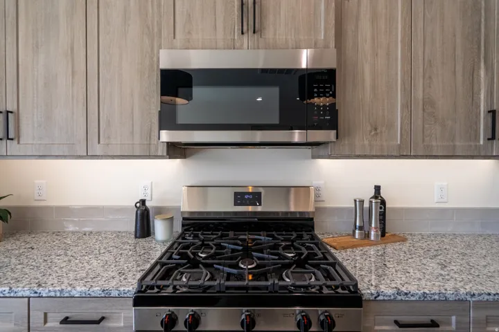 Contemporary kitchen featuring a five-burner gas cooktop with stainless steel finish, quartz stone countertops, stainless steel over-the-range microwave, soft-close cabinetry in warm wood tones, and under-cabinet lighting throughout