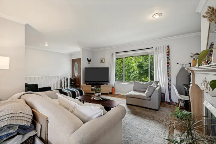 Living area featuring wood-finish flooring, a decorative fireplace, crown molding, and a large window overlooking lush greenery