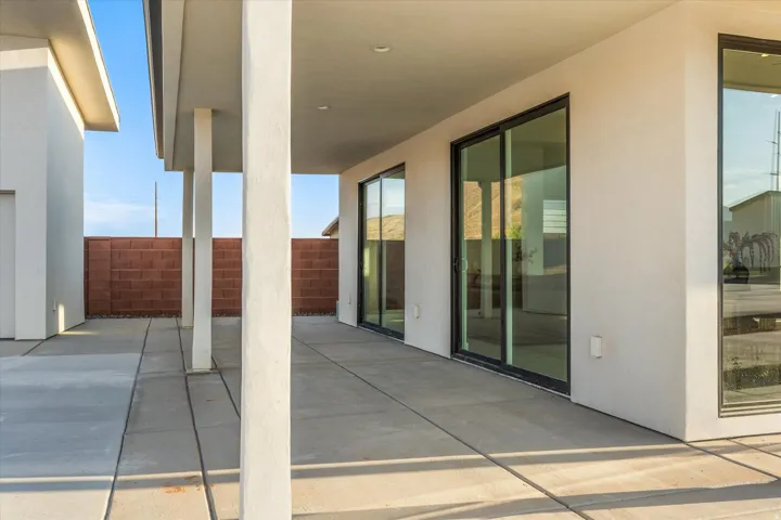 Expansive concrete patio with a covered outdoor area, supported by columns, featuring two sets of sliding glass doors with black frames, and a perimeter block wall