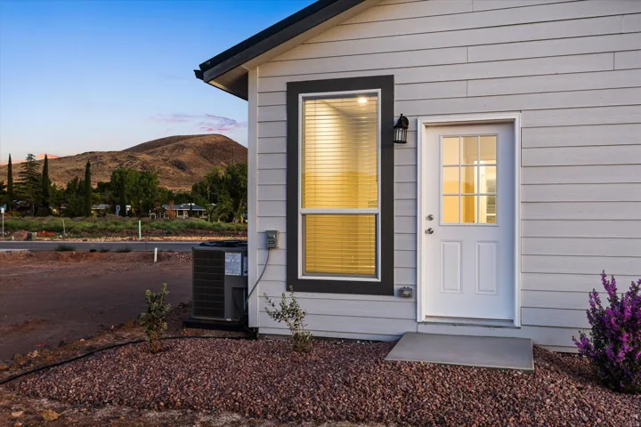 Light-colored horizontal siding, dark-trimmed window, white entry door with glass panels, exterior wall lantern, and a concrete entry pad