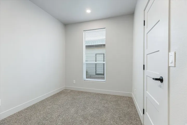 Neutral wall paint, carpet flooring, a window with horizontal blinds, a ceiling-mounted recessed light, and a white interior door with black hardware