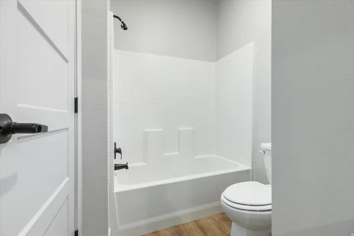 White paneled door with matte black hardware, white tub-shower combination with subway tile surround, matte black shower fixtures, and wood-finish flooring