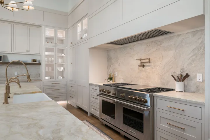 Gourmet kitchen with stainless steel range, full-height stone slab backsplash, pot filler, custom white cabinetry, and glass-front upper cabinets