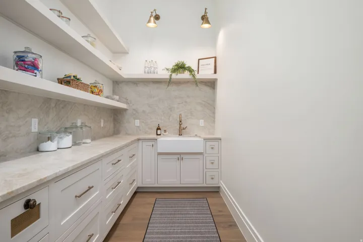 Built-in cabinetry with ample storage, integrated farmhouse sink with brass-finish faucet, stone slab countertops and backsplash, floating open shelving, and wood-finish flooring