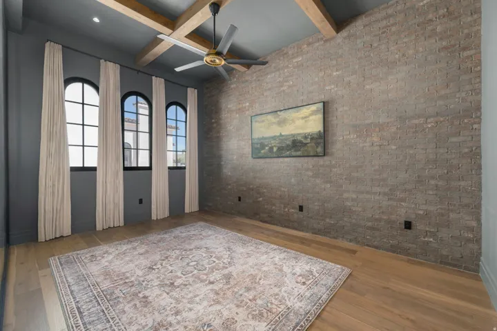 Hardwood flooring, exposed brick wall, three arched windows, ceiling fan, and coffered ceiling with wood beams