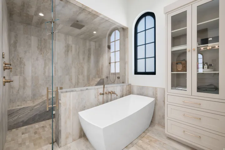 Glass-enclosed shower with integrated bench seating, freestanding soaking tub, arched window, and built-in cabinetry with glass display doors