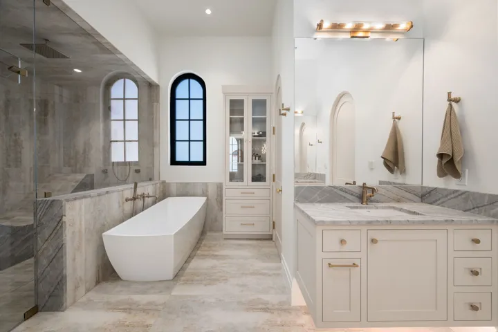 Freestanding soaking tub, walk-in shower with bench seating, dual arched windows, built-in storage cabinetry, and a vanity with stone countertop