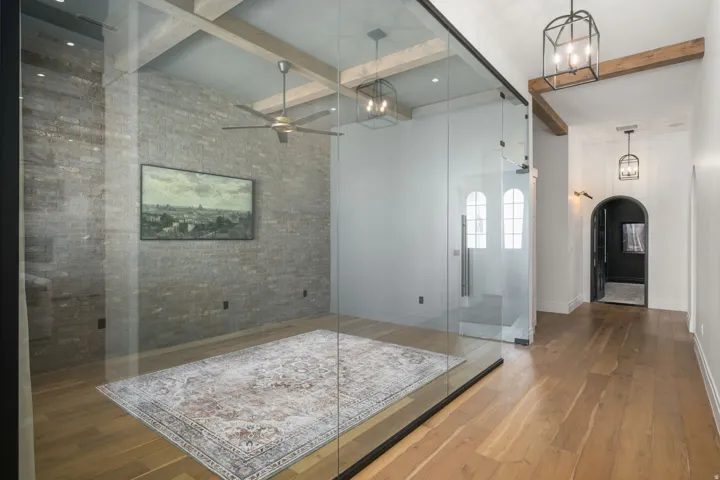 Wood-finish flooring throughout, exposed brick accent wall, ceiling fan, glass partition with a sliding door, and vaulted ceiling with exposed wood beams