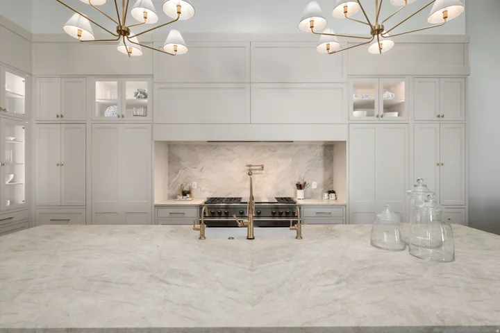 Expansive kitchen island with a light-toned countertop, custom cabinetry, built-in range, and dual brass chandeliers