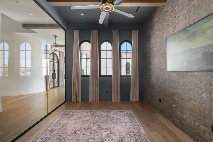 Wood-finish flooring, exposed brick accent wall, ceiling fan with light fixture, decorative wood beam ceiling, and arched windows