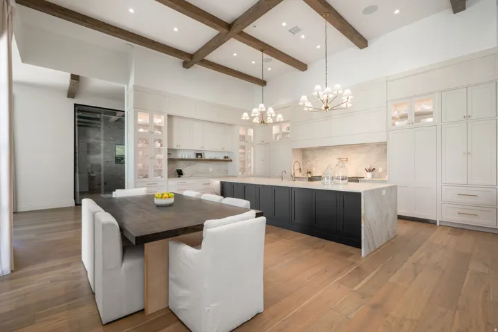Vaulted ceilings with exposed wood beams, wood-finish flooring, two-tone kitchen cabinetry, an oversized kitchen island with waterfall countertop, and multiple chandeliers