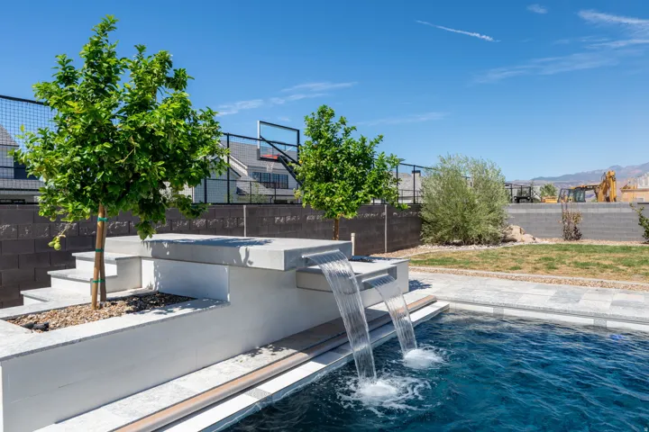 Swimming pool with integrated waterfalls, hardscape patio, and stone steps, surrounded by a block wall fence and mature trees