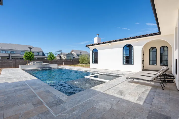 Outdoor swimming pool with integrated spa and waterfall features, surrounded by a spacious stone patio, set against a stucco exterior with arched windows