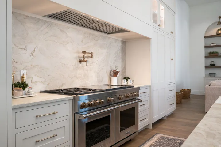 Gourmet kitchen with a professional-grade stainless steel range, full-height stone slab backsplash, pot filler faucet, custom cabinetry, and wood-finish flooring