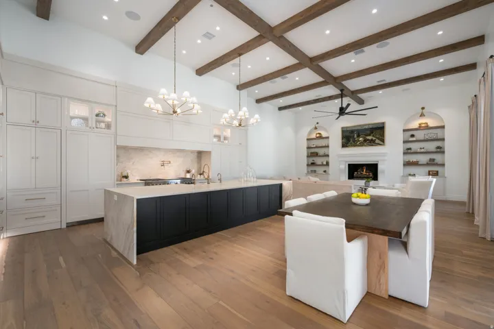 Spacious kitchen and living area featuring wood-finish flooring, exposed wood ceiling beams, white cabinetry, a large kitchen island with counter seating, and a fireplace