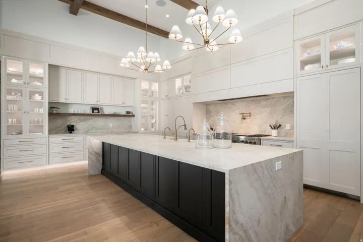 Spacious kitchen featuring an expansive island with waterfall countertops, wood-finish flooring, custom white cabinetry, and exposed ceiling beams