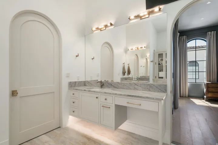 Arched doorway and entryway, expansive vanity with a light-toned countertop, integrated dual sinks with bronze-toned fixtures, and a large wall-mounted mirror with overhead lighting