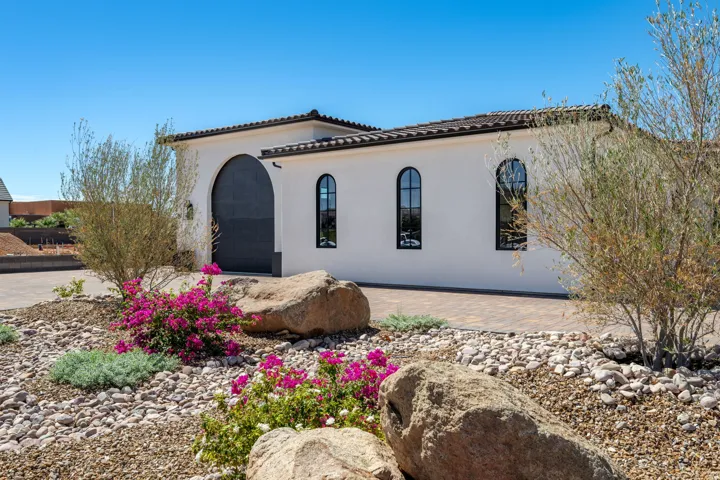 Stucco exterior with arched windows, tile roofing, paver driveway, and a xeriscape landscape featuring mature plantings and decorative boulders