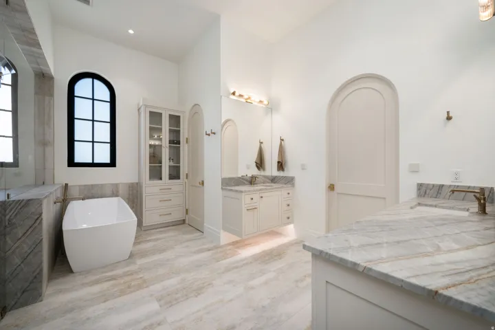 Freestanding soaking tub, arched window, dual vanity with stone countertops, built-in storage cabinet, and wood-finish flooring