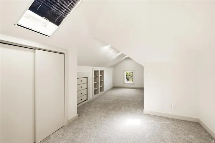 Vaulted ceiling room with a skylight, soft gray carpet, and a built-in storage unit with shelving and drawers