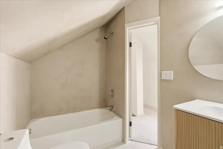 Bathtub with chrome shower fixtures, round vanity mirror, and slatted wood-finish vanity with an integrated sink
