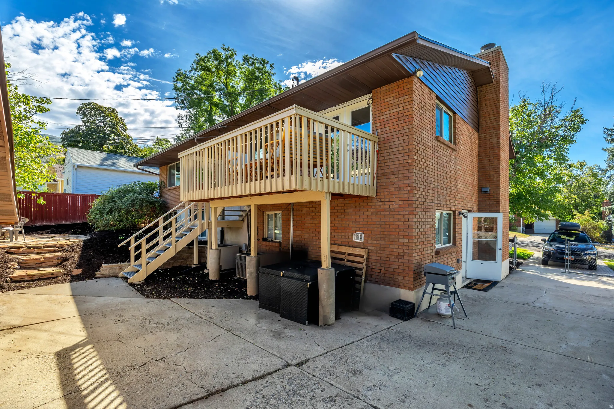 Brick exterior with an elevated wood deck and staircase, concrete patio, and landscaped planting beds