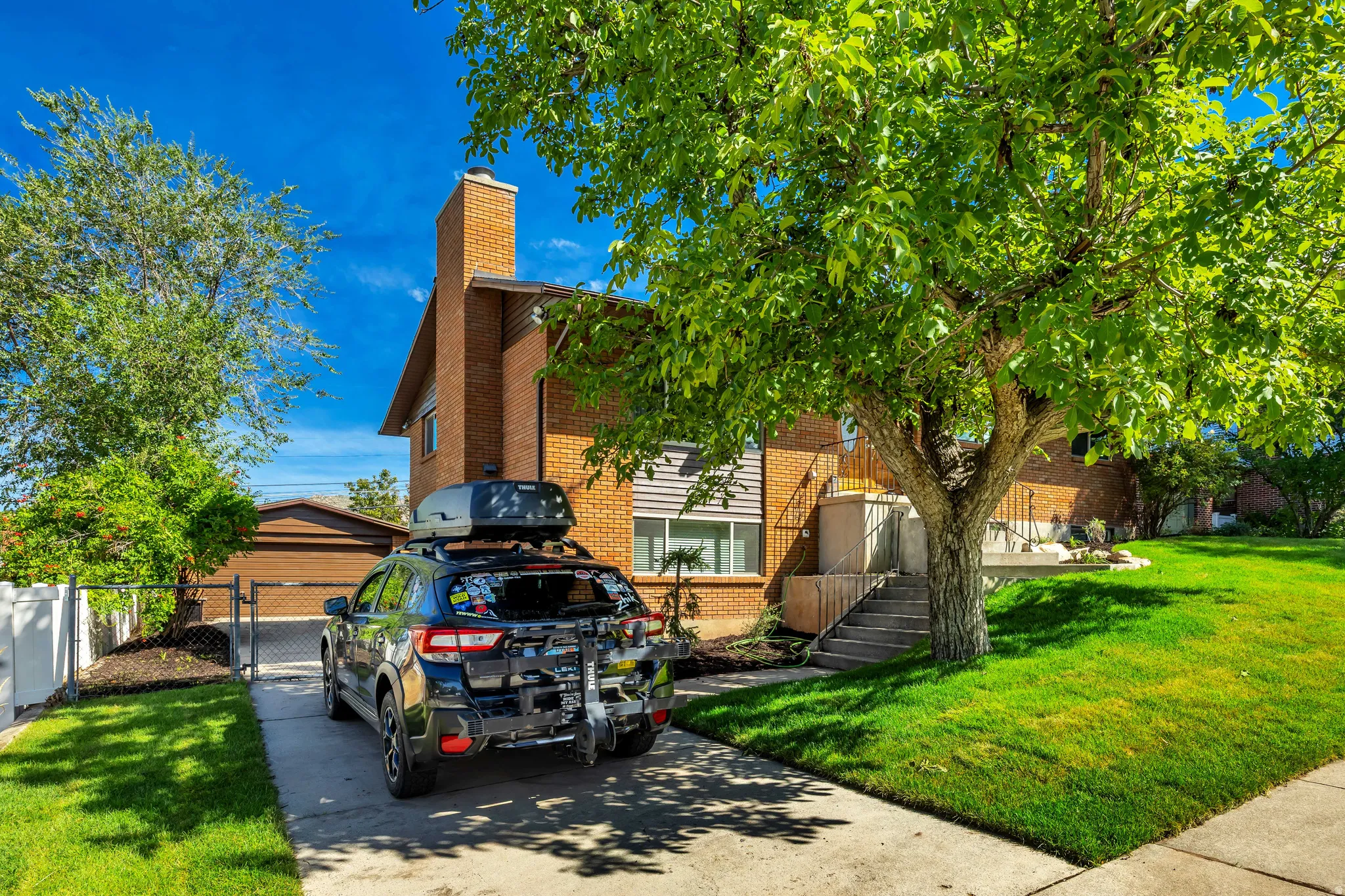 Brick exterior with a prominent chimney, concrete driveway, attached garage, mature trees, and a sloped lawn