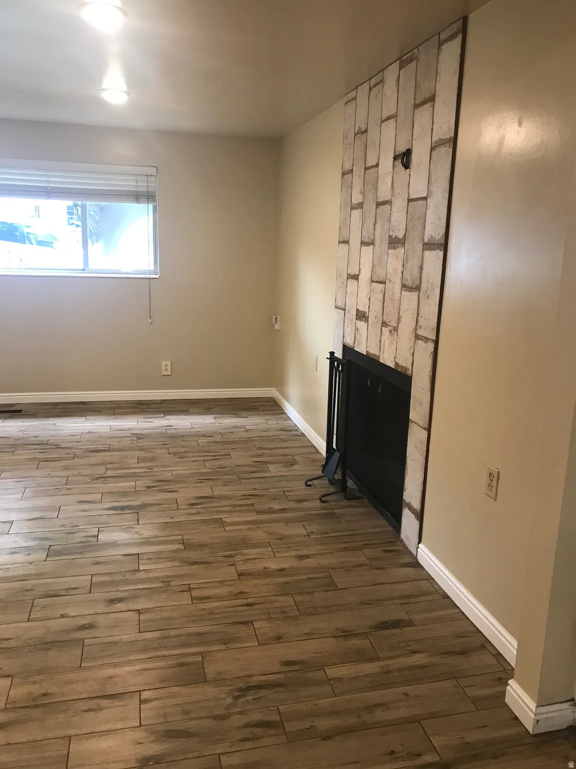 Wood-finish flooring, white brick-style fireplace surround, black fireplace screen, recessed lighting, and a window with blinds
