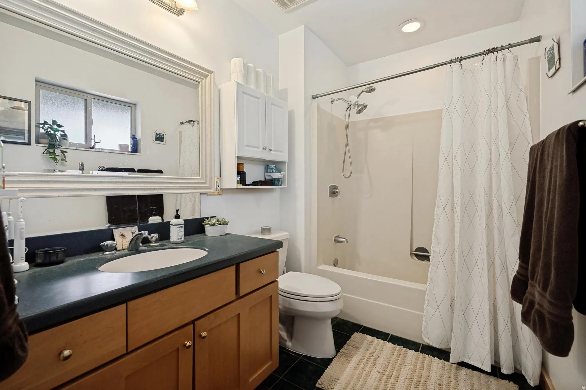 Bathroom featuring a vanity with wood-finish cabinetry, an integrated sink, a large framed mirror, a tub-shower combination, and dark tile flooring