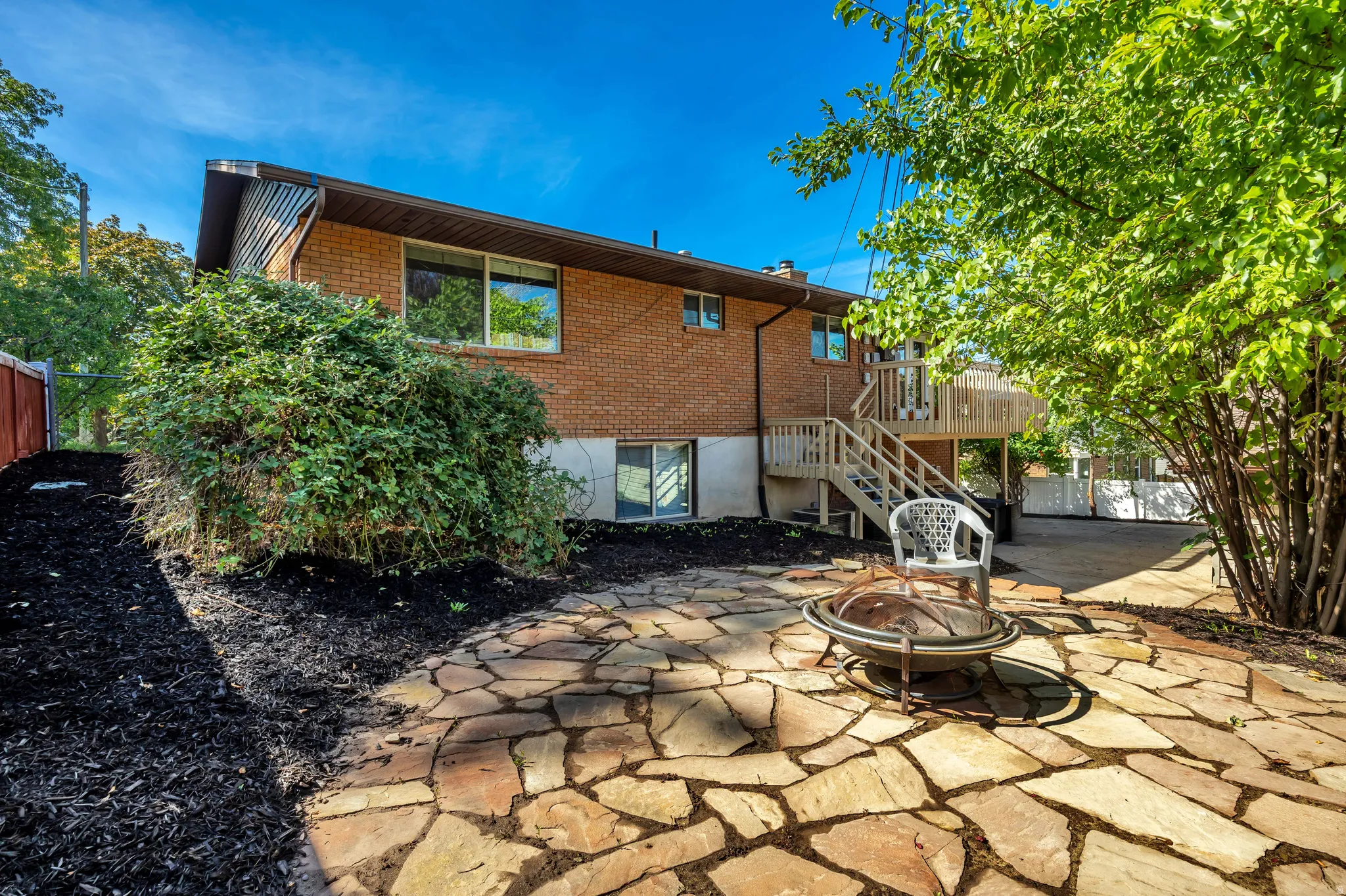 Brick exterior with multiple windows, leading to an expansive flagstone patio, a wood deck with stairs, and a fire pit