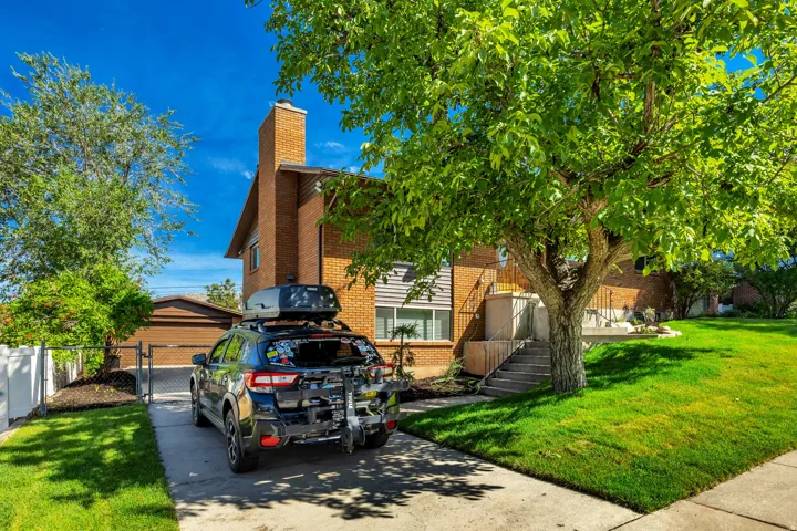 Brick exterior with a prominent chimney, concrete driveway, attached garage, mature trees, and a sloped lawn