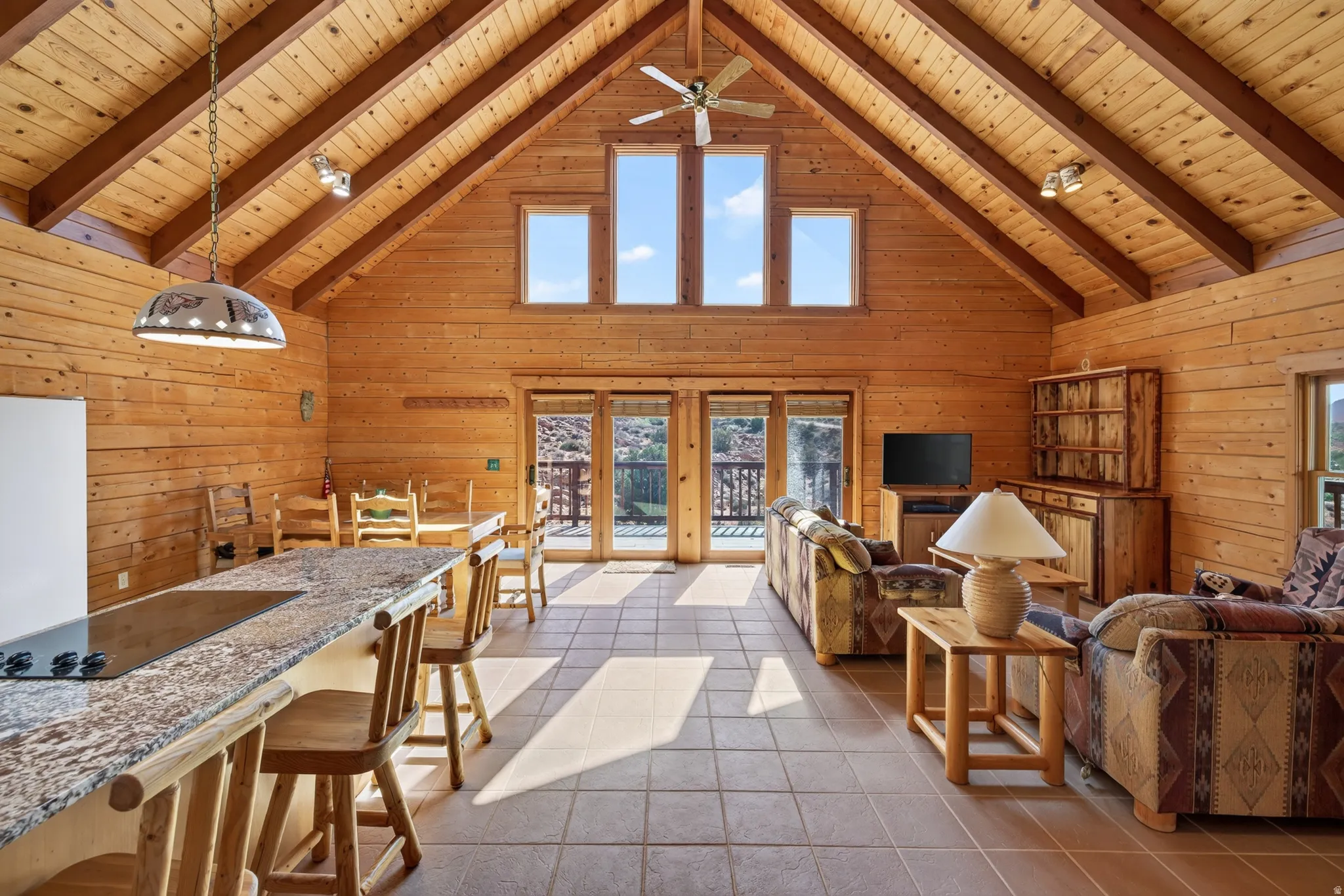 Cathedral ceiling with exposed wood beams, extensive natural wood wall paneling, full-height windows, tile flooring, and integrated kitchen island with cooktop