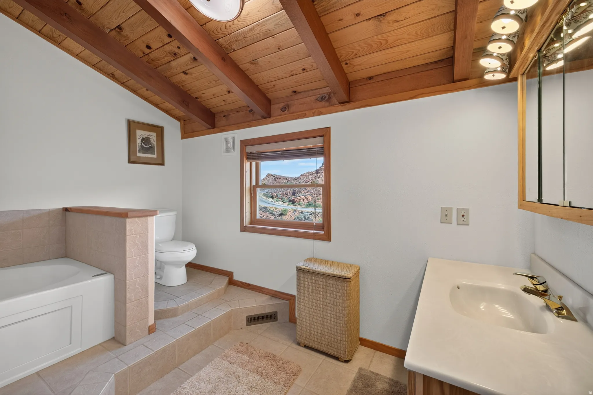 Vaulted wood-plank ceiling with exposed beams, wood-framed window, built-in bathtub with tiled surround, and a vanity with a drop-in sink