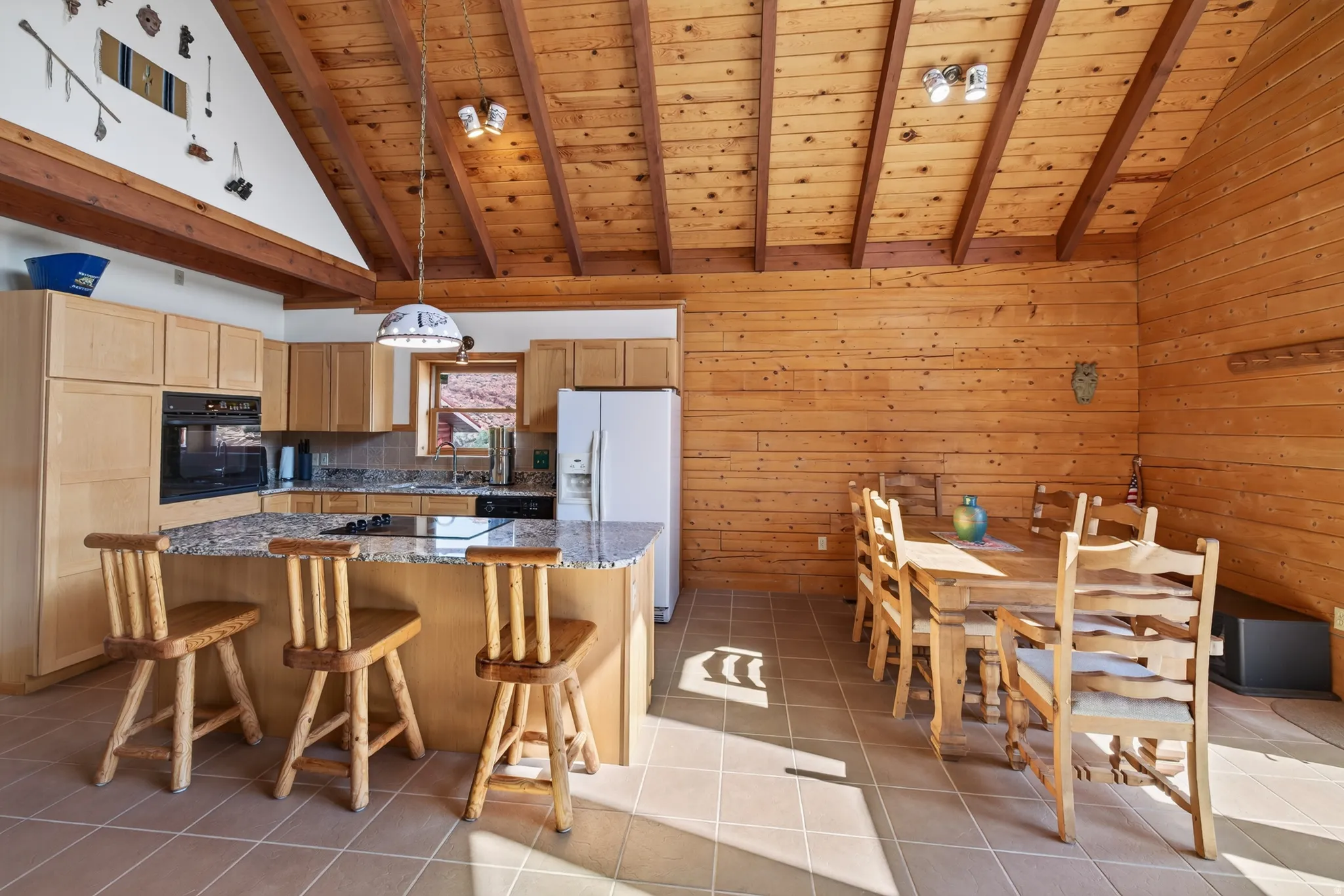 Vaulted wood ceiling with exposed beams, wood-paneled walls, tile flooring, kitchen island with built-in cooktop, and light wood cabinetry