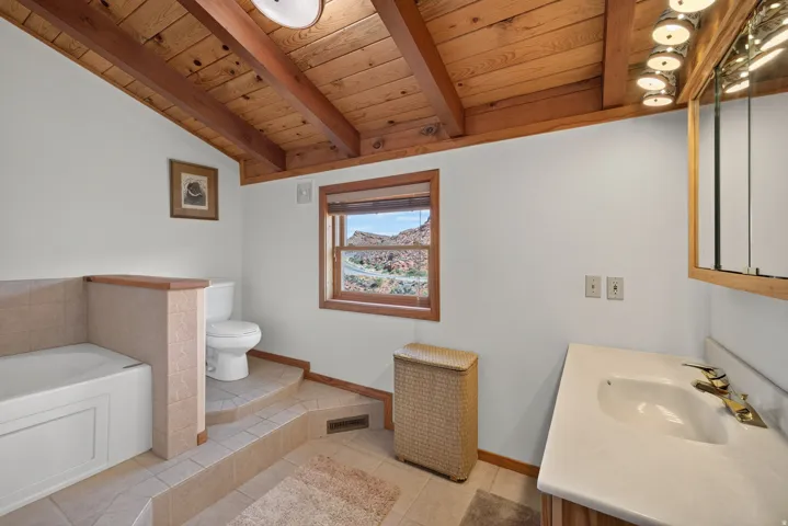 Vaulted wood-plank ceiling with exposed beams, wood-framed window, built-in bathtub with tiled surround, and a vanity with a drop-in sink