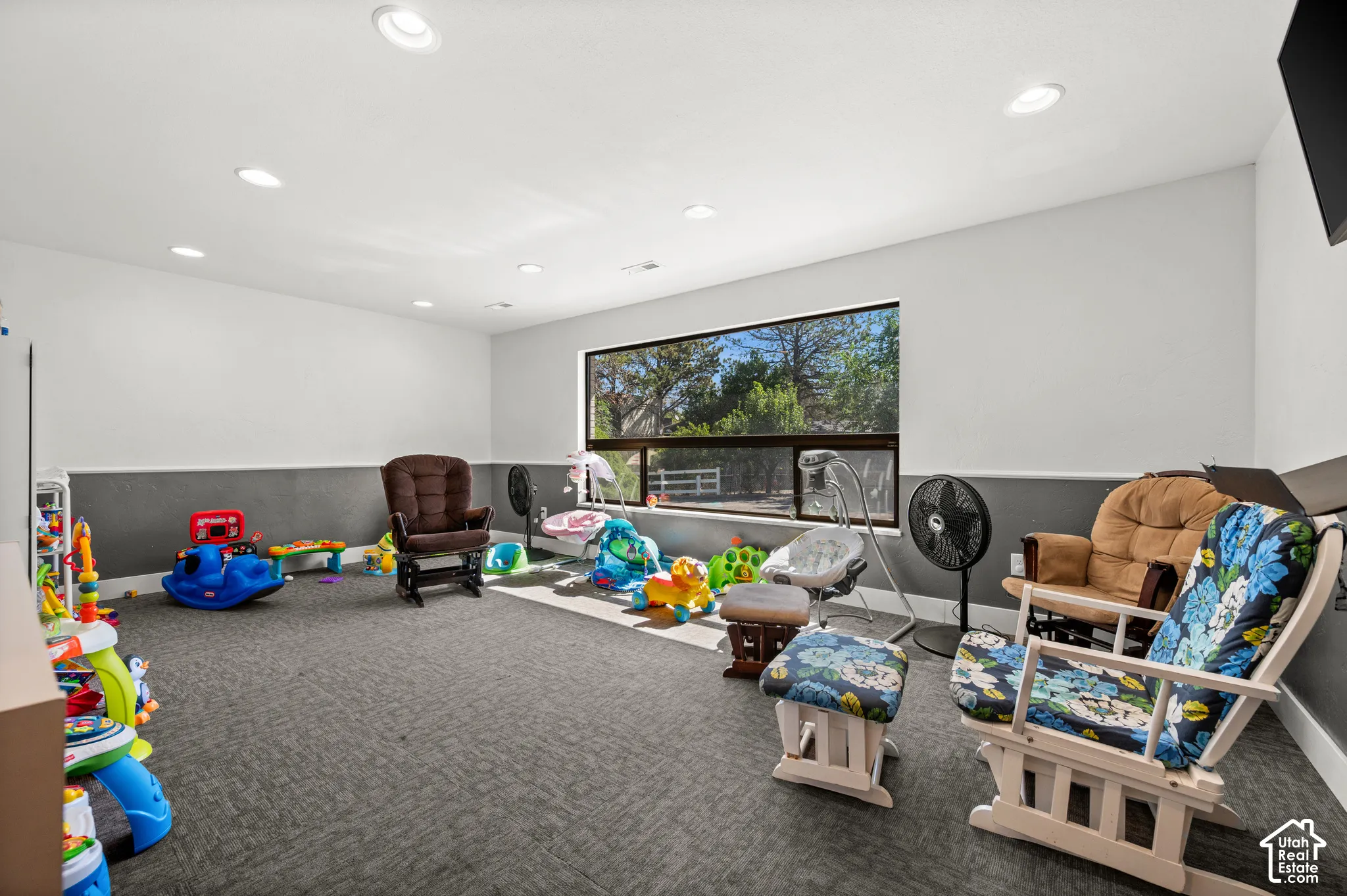 Recessed ceiling lighting illuminates a spacious room featuring a large window with a roller shade, and a textured carpeted floor