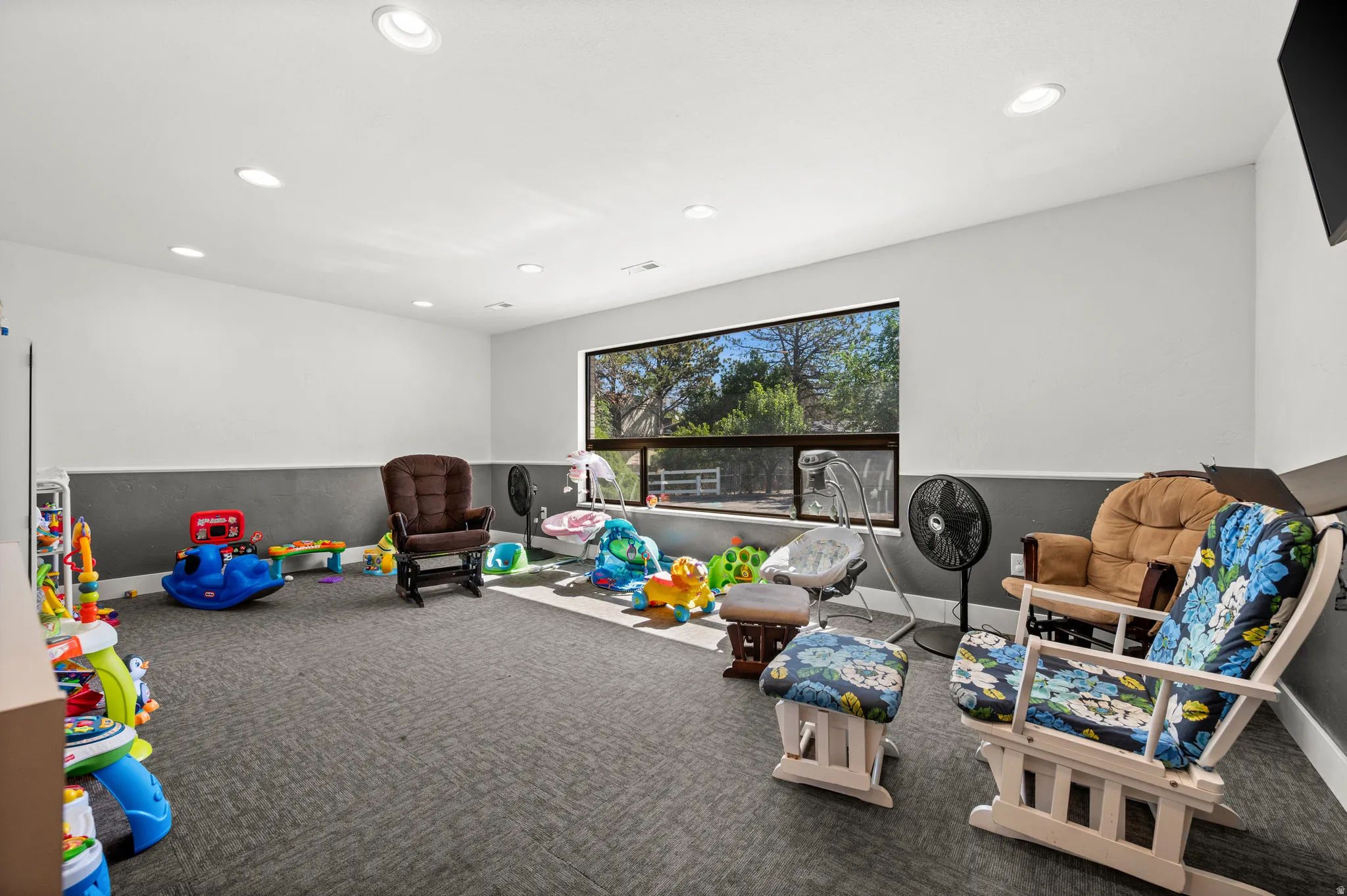 Recessed ceiling lighting illuminates a spacious room featuring a large window with a roller shade, and a textured carpeted floor
